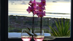 A window with a view of the sea and a table with two glasses of wine and purple orchids.