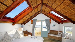 A cozy bedroom with a sloped ceiling, a bed with white linens, a wicker chair, a small table with a lamp, and a view of the sea through a window. - Gilcrest Place (Paternoster)