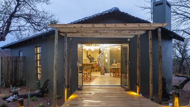 Um deck de madeira leva a uma casa com entrada coberta, área de refeições interna e uma grande árvore no exterior.