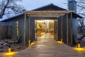 Una terraza de madera que lleva a una casa con un porche cubierto, un comedor en el interior y un gran árbol afuera.
