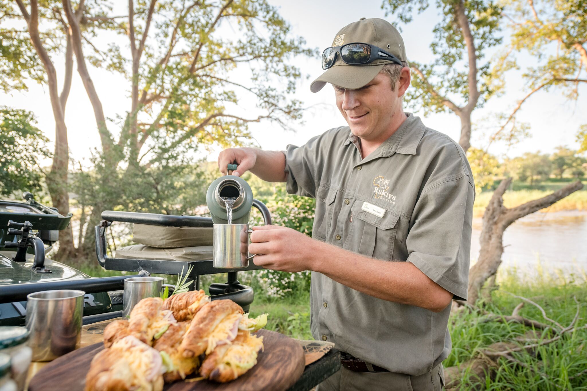A person pouring a drink from a kettle onto a mug, with a table of pastries in the foreground and a natural outdoor setting in the background.