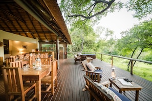 A wooden deck with tables and chairs, a thatched roof, and a view of greenery.