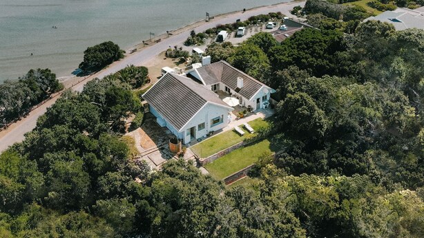 Eine Luftaufnahme einer Küstenimmobilie mit einem großen Haus, einer privaten Zufahrt und einem Strand.