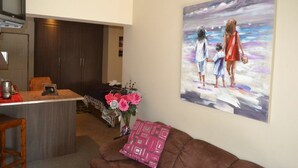 A living room with a brown sofa, a painting of a family on a beach, a kitchen area with a TV, and a small table with a vase of flowers. - Ikhanda Guesthouse (Mashishing)