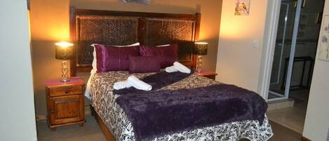 Een net opgemaakt bed met een beddekleed met een patroon, twee kussens en een paarse plaid. Er staan twee nachtlampjes op houten nachtkastjes.