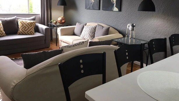A modern living room with a sofa, dining table, and chairs. There are two paintings on the wall and a lamp on the table. - Stell On Air (Stellenbosch)