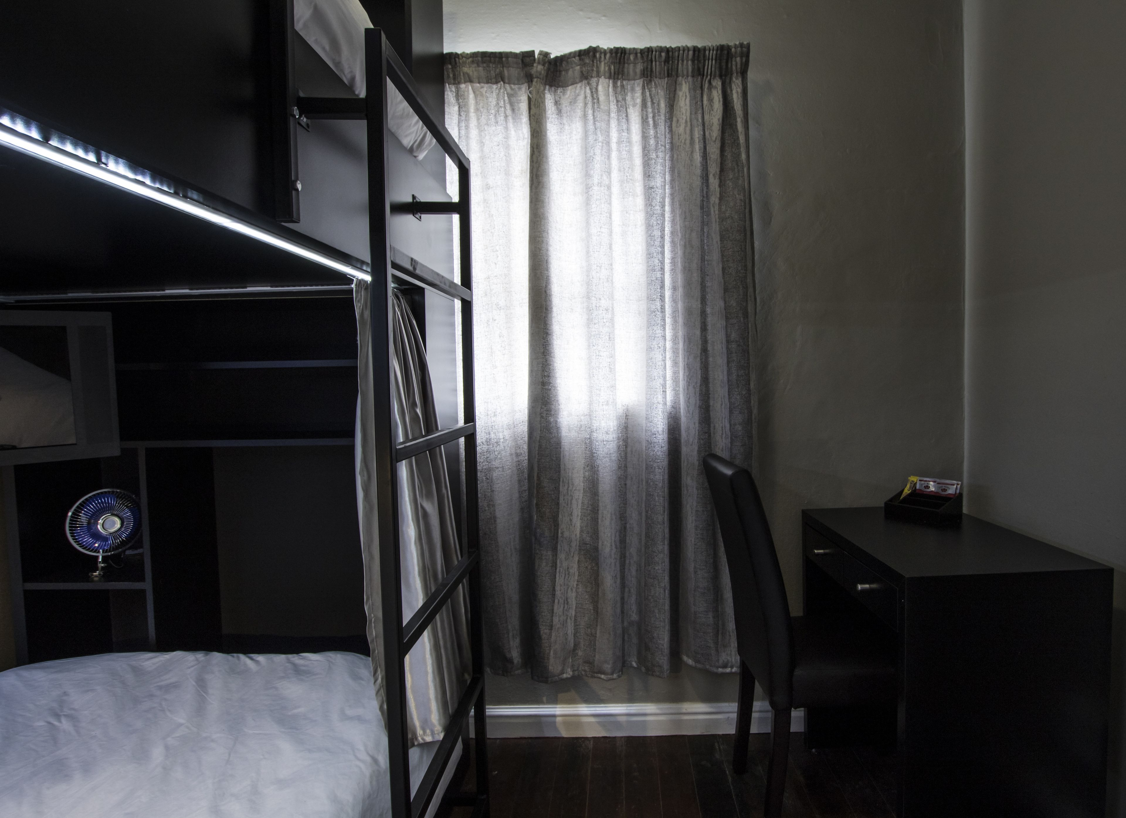 Quarto com beliche, escrivaninha, cadeira, ventilador e janela com cortinas.