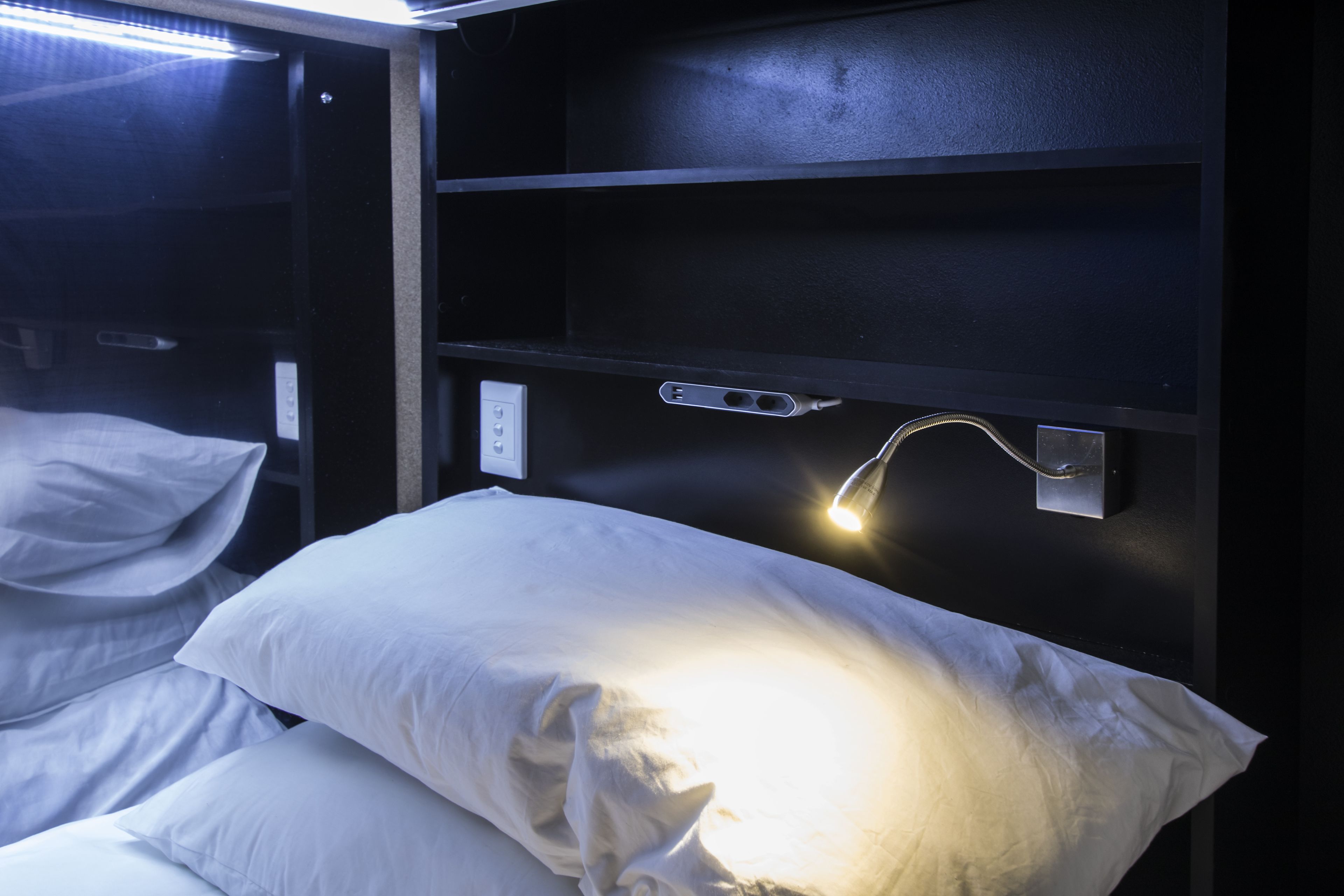 A dimly lit room with a single bed, white bedding, and a wall-mounted light fixture.