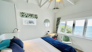 A bedroom with a bed, a desk, a chair, a window with a view of the sea, and a ceiling fan. - Sunny Cove Manor (Cape Town)