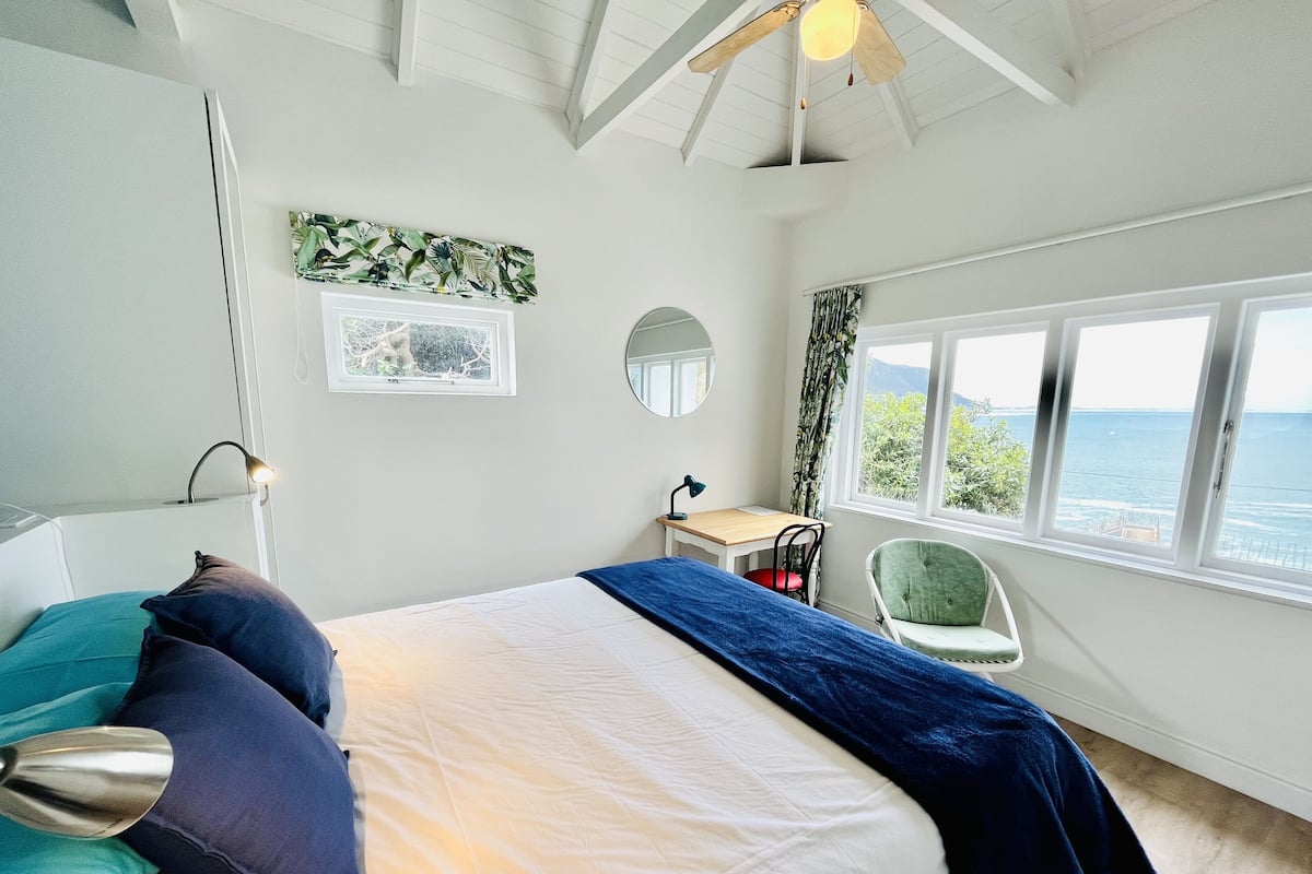 Un dormitorio con cama, escritorio, silla, ventana con vista al mar y ventilador de techo.