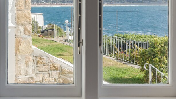 A view from a window overlooking a coastal landscape with a stone wall, a grassy area, and a metal railing. - Sunny Cove Manor (Cape Town)