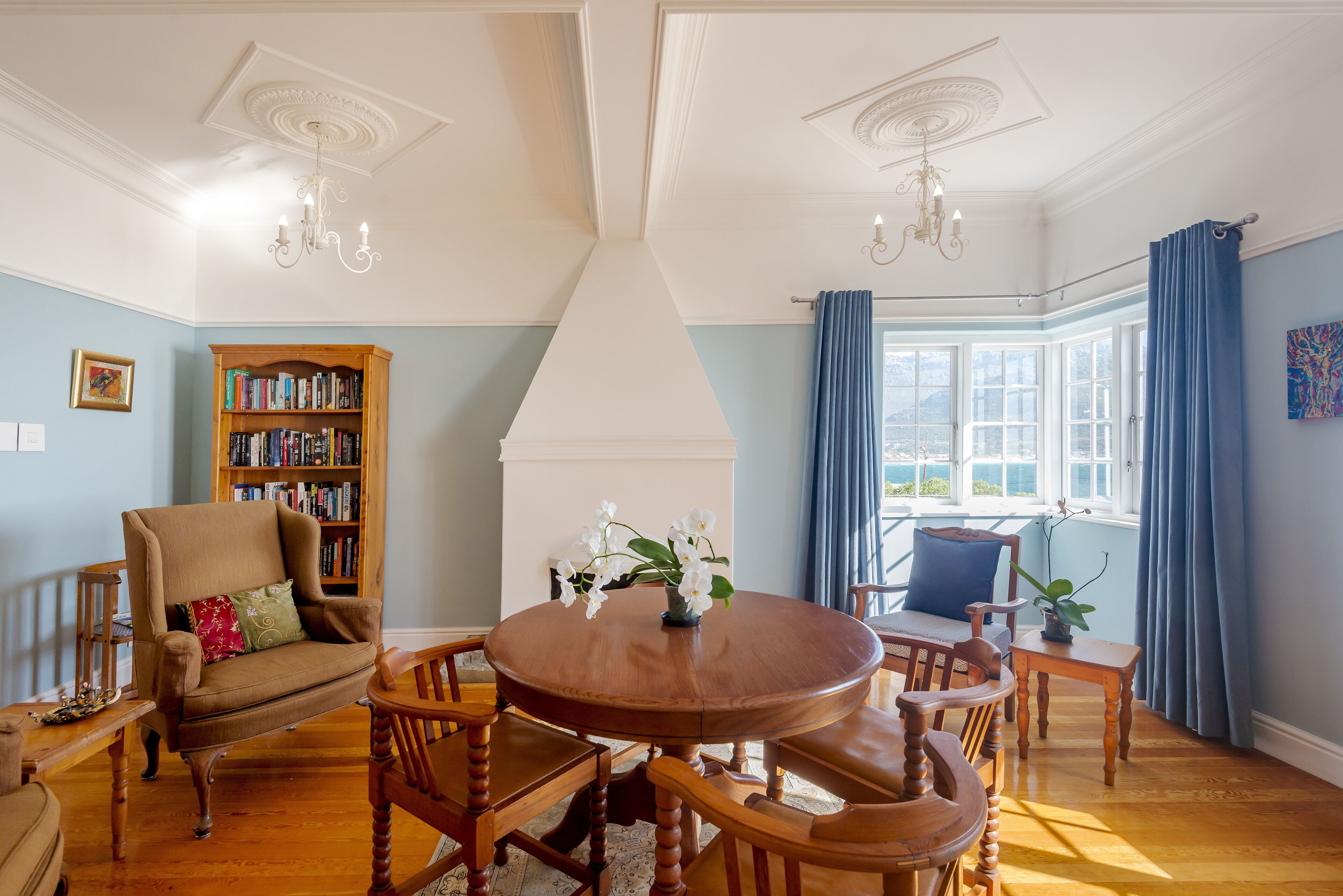 A dining area with a round wooden table, chairs, a bookshelf, and a window with curtains.