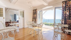 A dining area with white metal chairs and a table, large windows with a view of the sea, and a balcony with a railing. - Sunny Cove Manor (Cape Town)