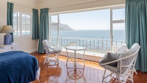 A room with a bed, a table, two chairs, and a view of the sea through large windows. - Sunny Cove Manor (Cape Town)