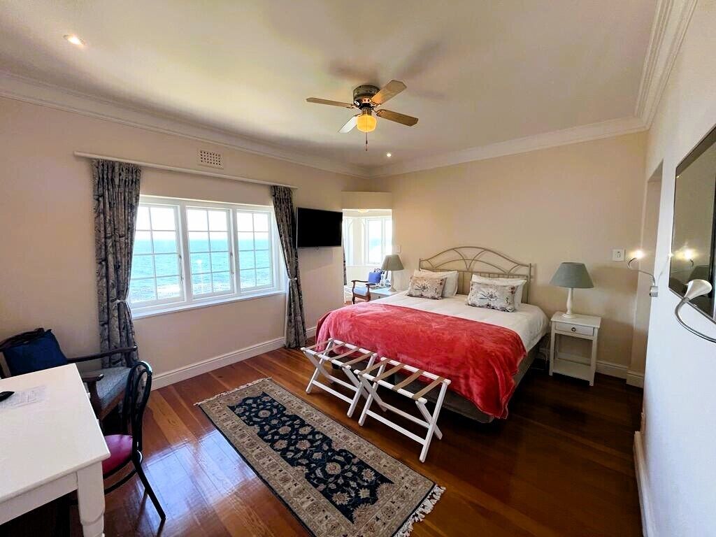 Une chambre à coucher comprenant un lit, des tables de chevet, un bureau, une chaise, un ventilateur de plafond et une vue sur la mer par une fenêtre.