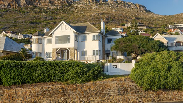 A large white house with a stone wall in the foreground, a number 72 sign, and a rocky hillside in the background. - Sunny Cove Manor (Cape Town)