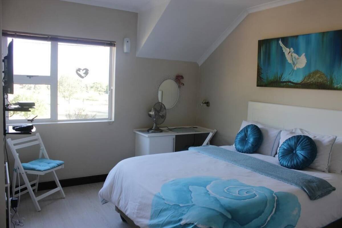 A bedroom with a bed, a desk, a chair, a window with a heart decoration, a painting, and a mirror.