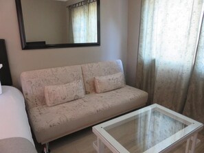 A room with a sofa, a glass-top coffee table, a mirror, and a window with curtains.