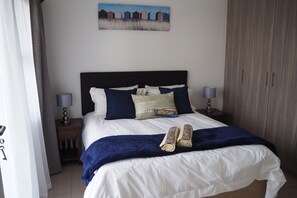 Una cama bien hecha con una manta azul, almohadas y una toalla enrollada. Hay dos mesitas de noche con lámparas, un cuadro en la pared y un armario.
