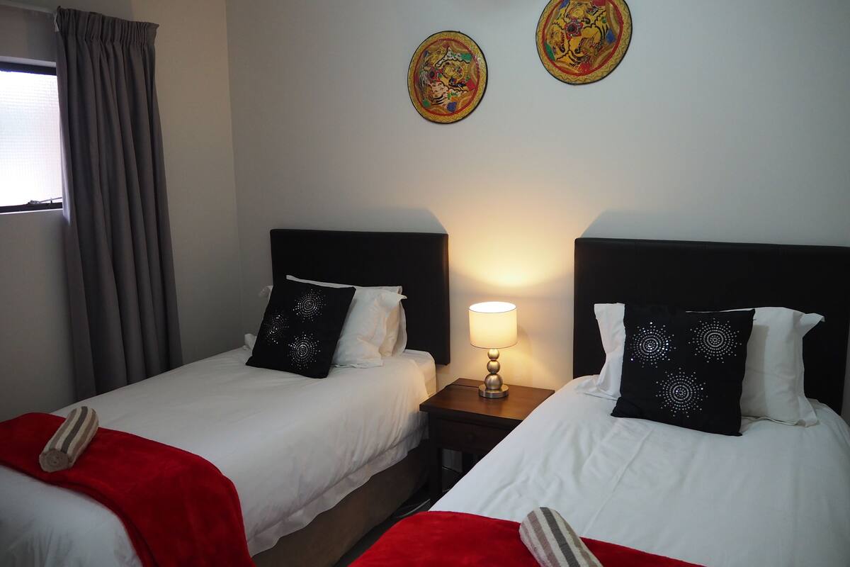 Dos camas individuales con sábanas blancas y almohadas decorativas negras, una mesita de noche con lámpara y una ventana con cortinas grises.