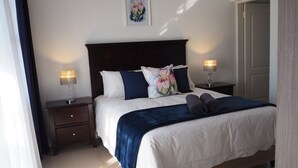 A neatly made bed with white and navy bedding, two bedside lamps, a dark wooden headboard, and a framed floral artwork on the wall.