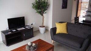 A modern living room with a flat-screen TV, a dark sofa with a yellow pillow, a wooden coffee table, and a potted plant.