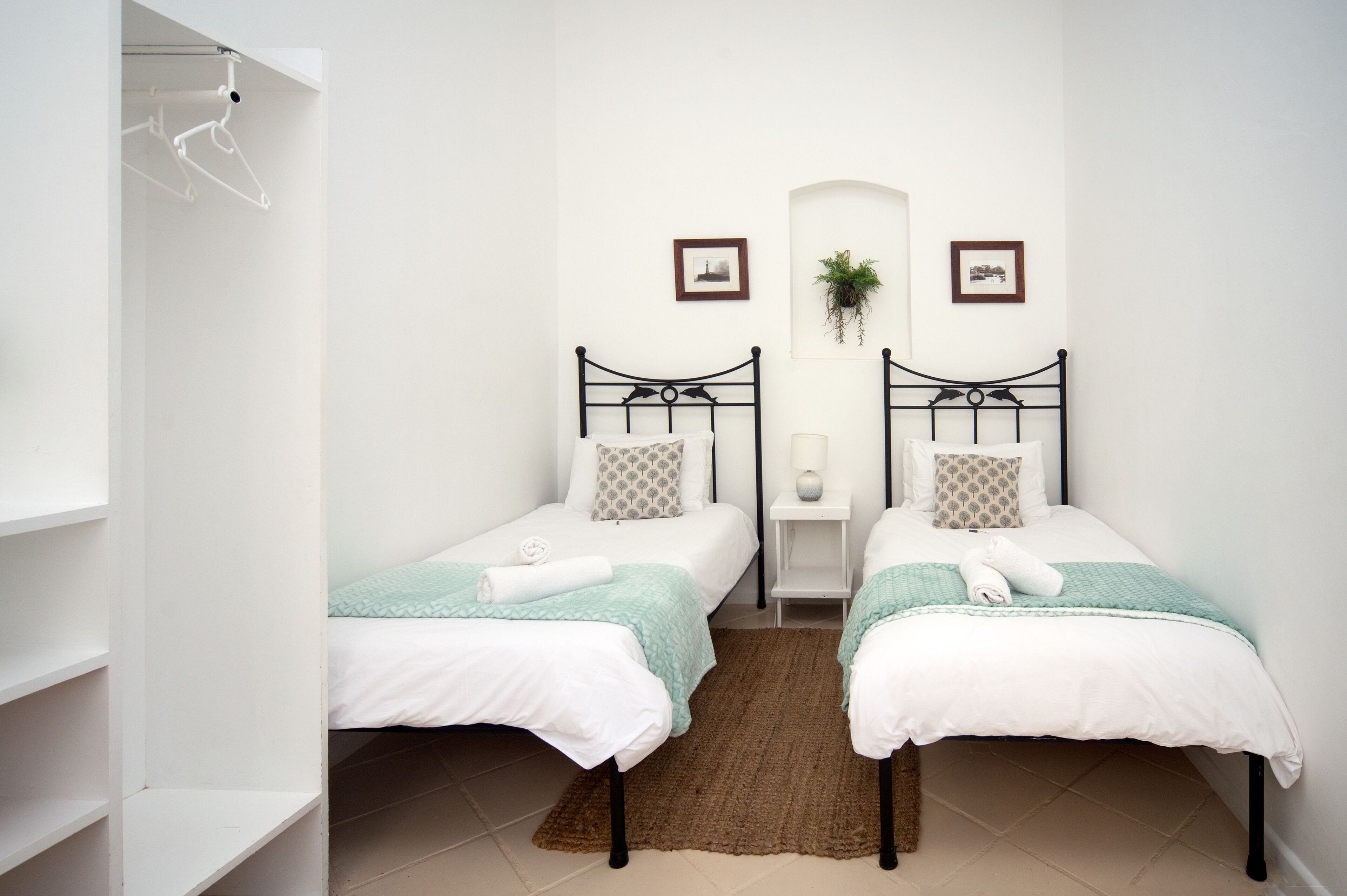 Camera da letto con due letti singoli, un piccolo comodino con lampada, una pianta appesa al muro e un quadro incorniciato.