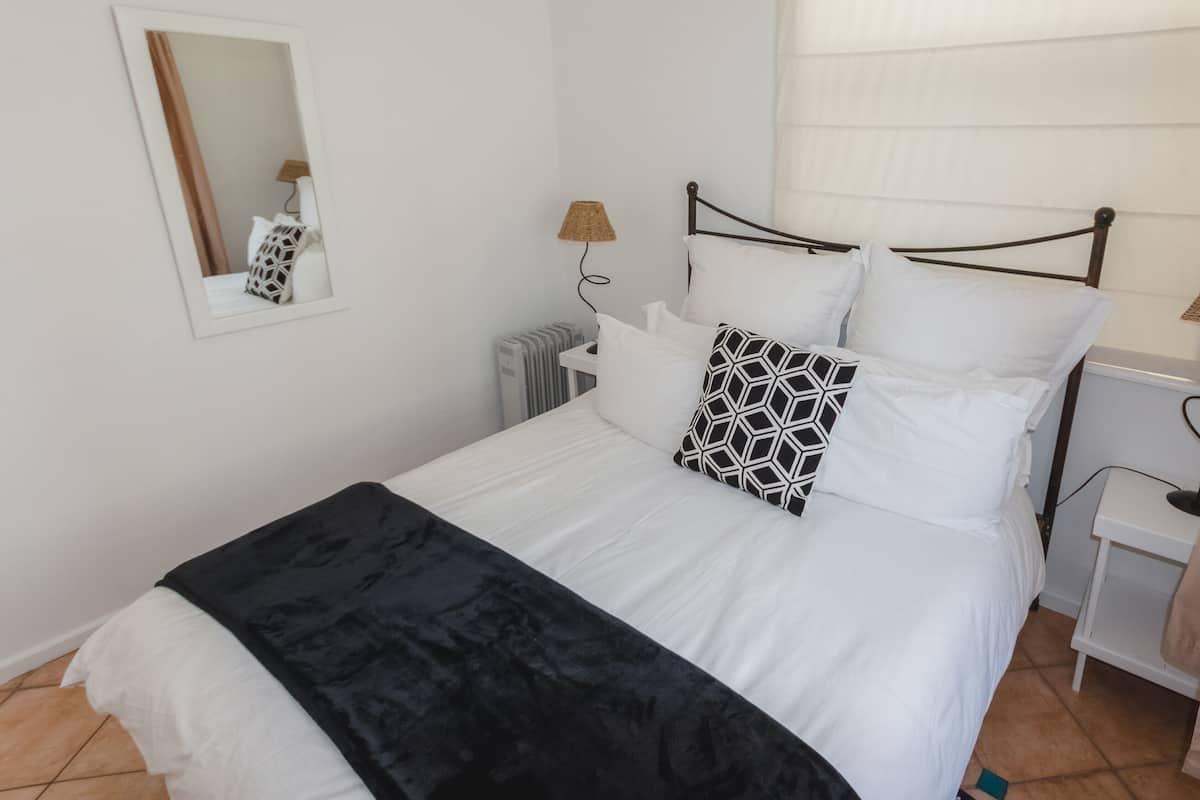 Ein ordentlich bezogenes Bett mit weißen Laken und einer schwarzen Tagesdecke, eine Nachttischlampe und ein Spiegel an der Wand.