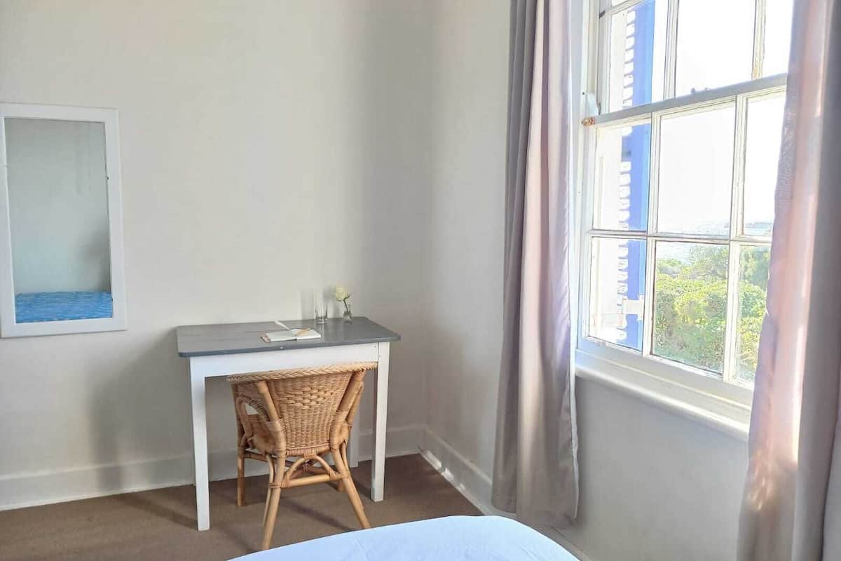 Ein Schlafzimmer mit Bett, Schreibtisch, Stuhl, einem Fenster mit Vorhängen und einem Wandspiegel.