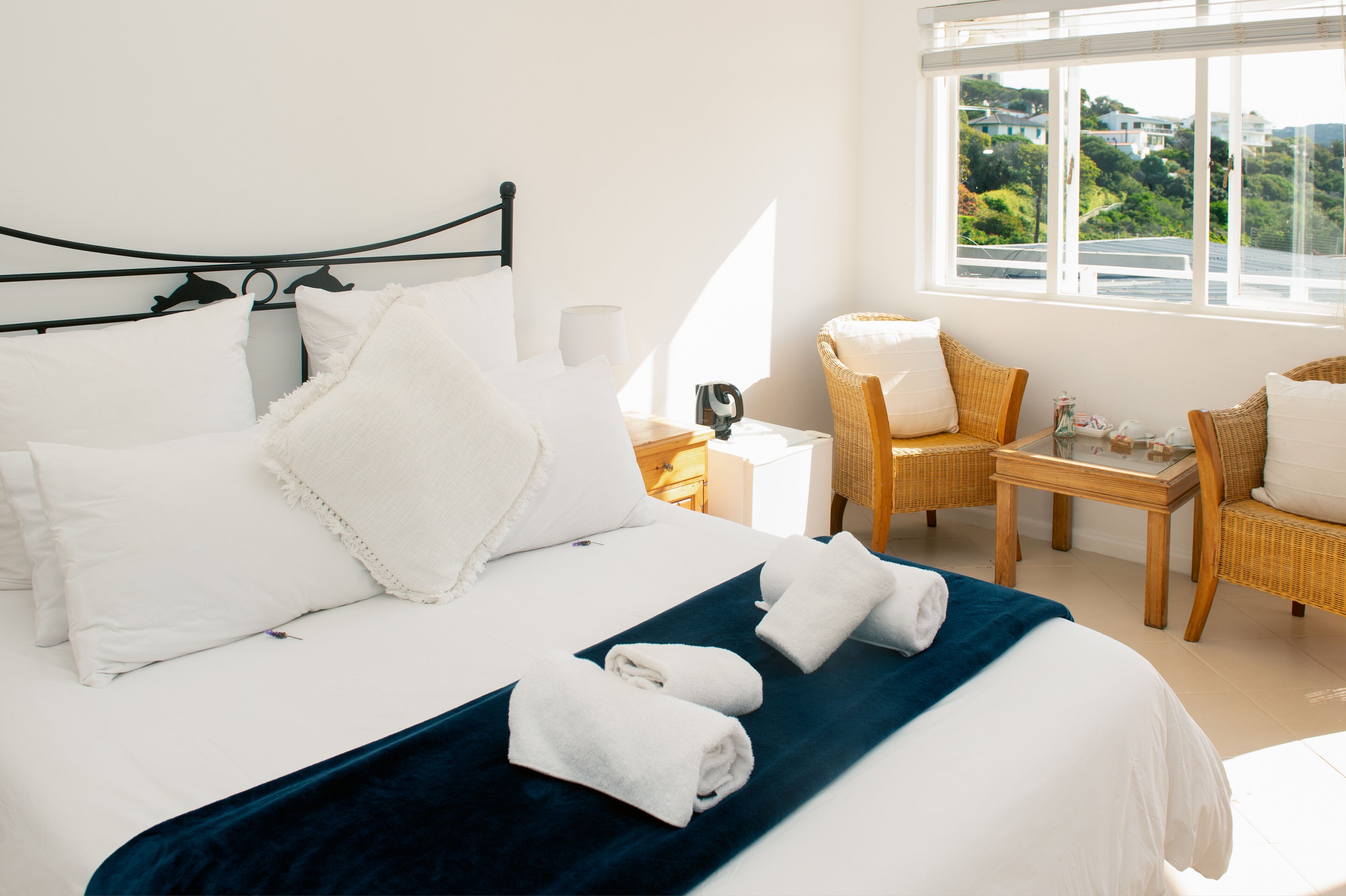 A neatly made bed with white linens and a blue accent, a wicker chair, and a small table with a lamp.
