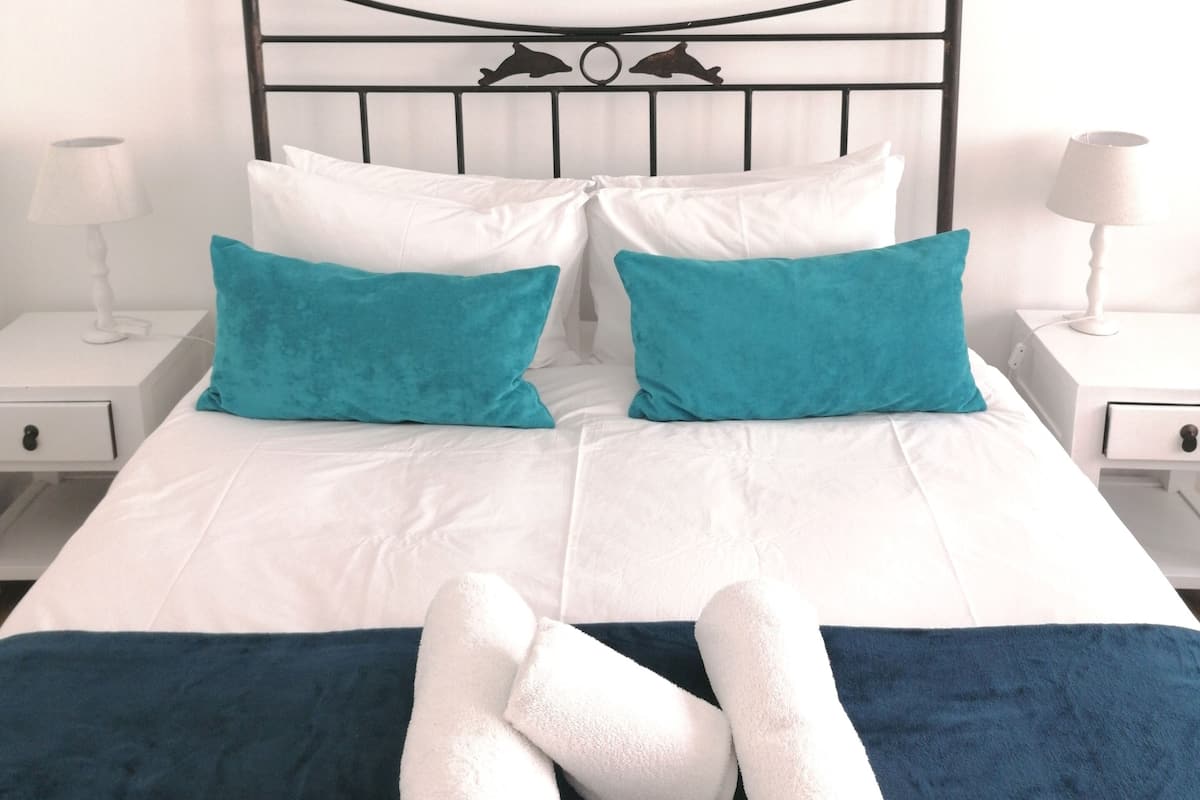 Ein ordentlich bezogenes Bett mit weißen und türkisfarbenen Kissen, einer türkisfarbenen Tagesdecke und aufgerollten weißen Handtüchern.