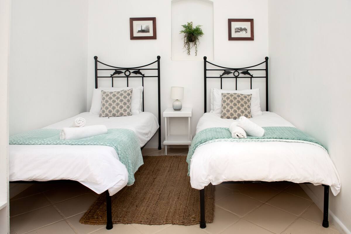 Ein Zimmer mit zwei Einzelbetten, einem kleinen Nachttisch mit Lampe, einem braunen Teppich und gerahmten Bildern an der Wand.