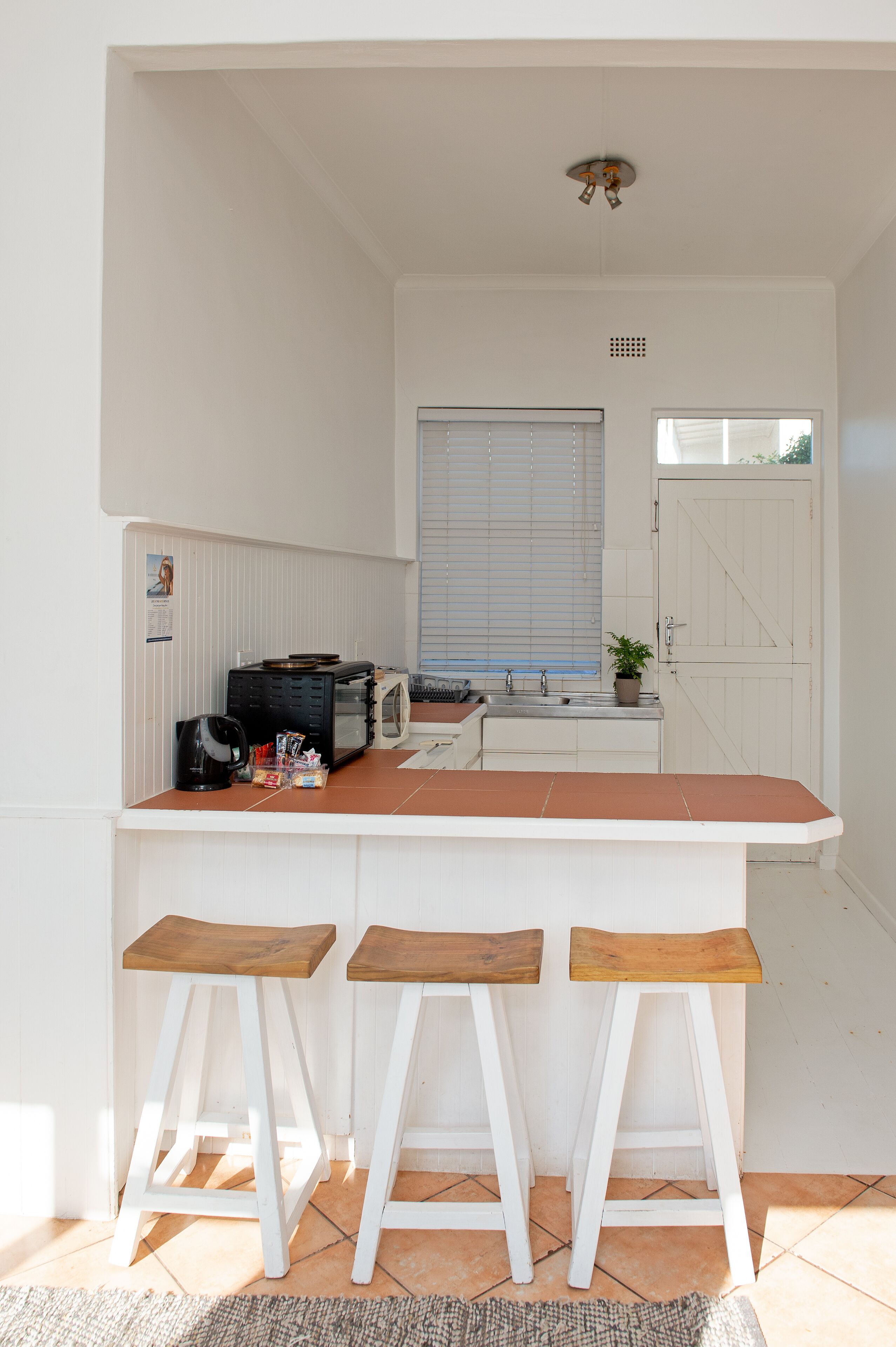 Cocina compacta con encimera blanca, tres taburetes de madera, un microondas, una tetera y una pequeña planta en maceta.