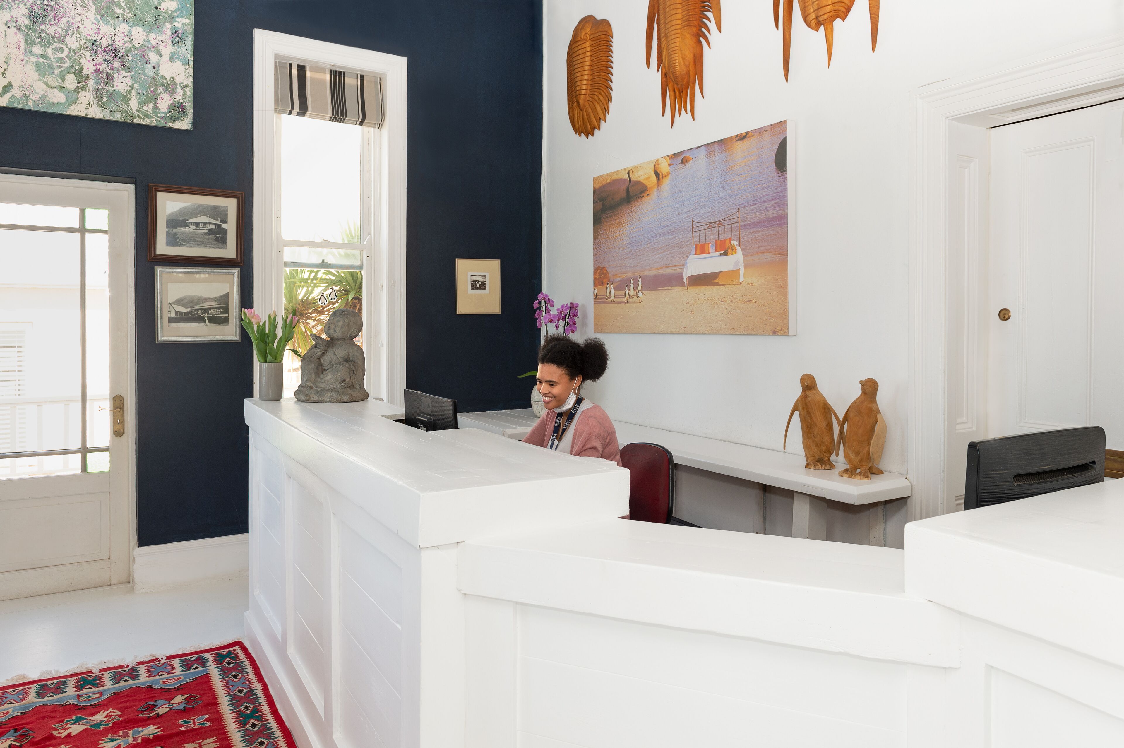 Un mostrador de recepción con una persona sentada detrás, una ventana con persianas y una pared decorada con cuadros y adornos.