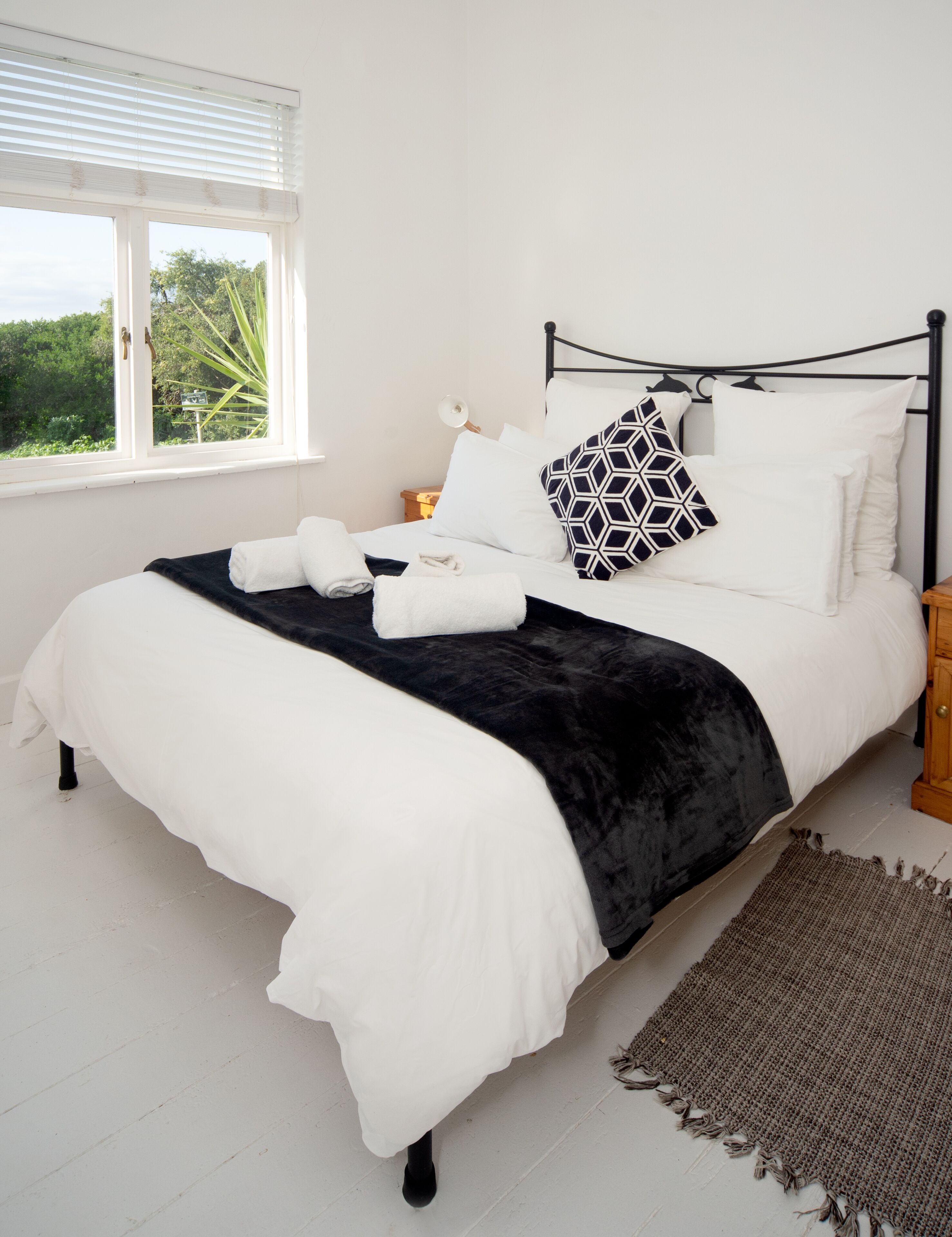A neatly made bed with white and black bedding, a bedside table with a lamp, and a view of greenery through a window.