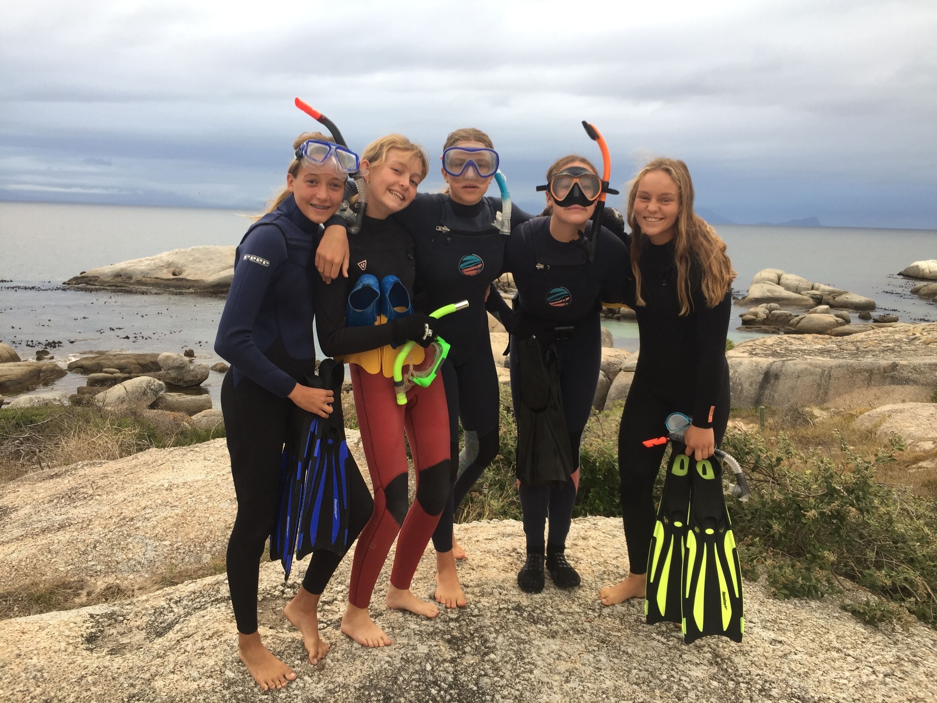 A group of people in wetsuits and snorkeling gear standing on a rocky shore with a body of water in the background.