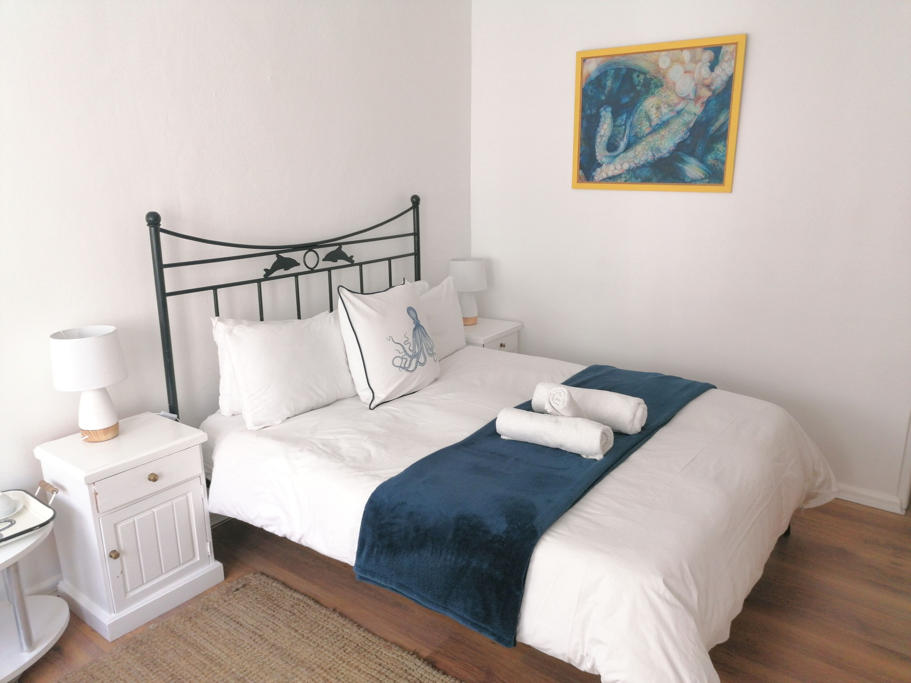 A neatly made bed with white bedding and a blue throw blanket, a bedside table with a lamp, and a wall-mounted painting.