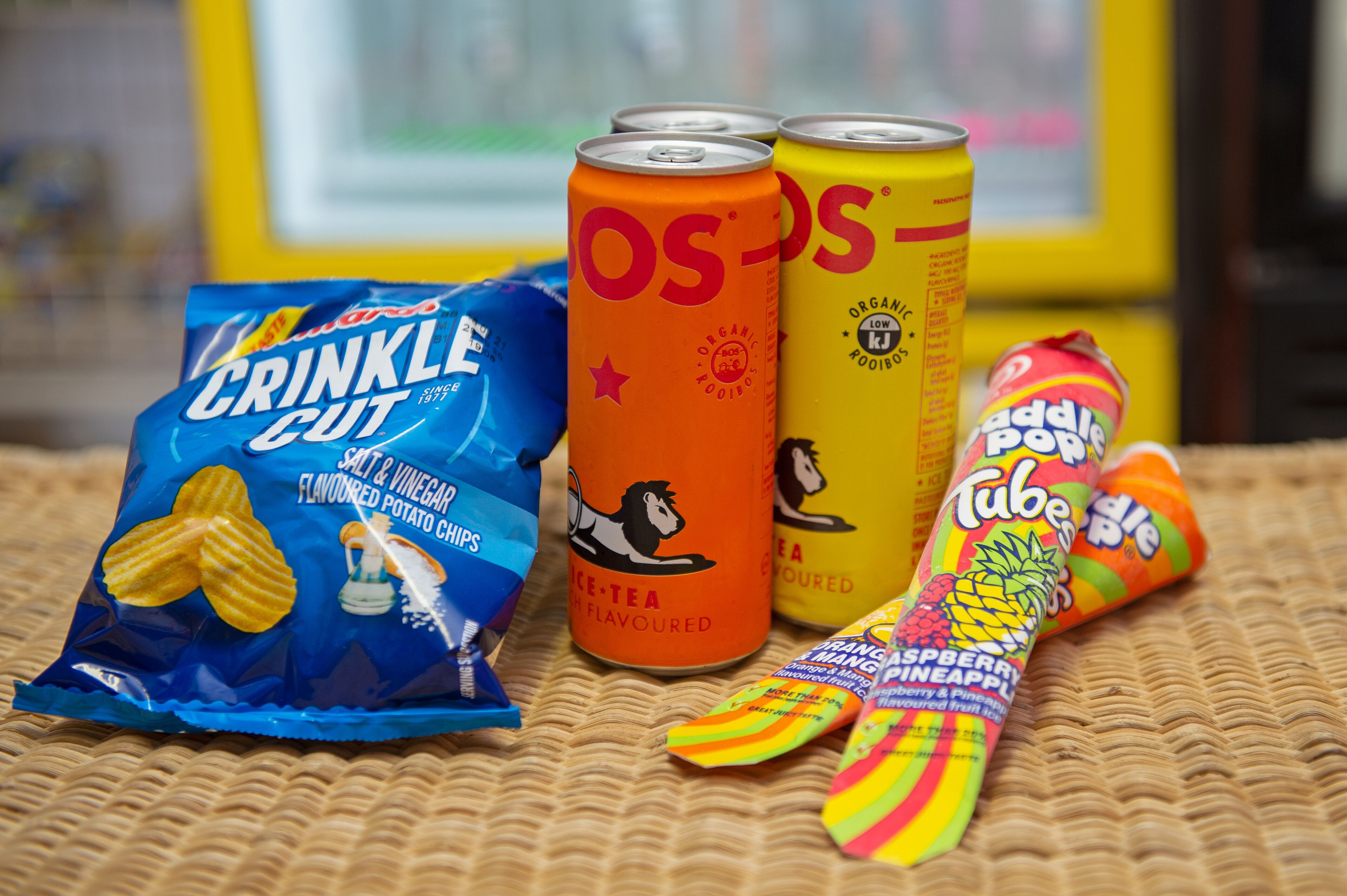 Un assortiment de collations comprenant des chips de pommes de terre à la coupe ondulée, deux canettes de thé glacé biologique et un tube de bonbons Paddle Pop.