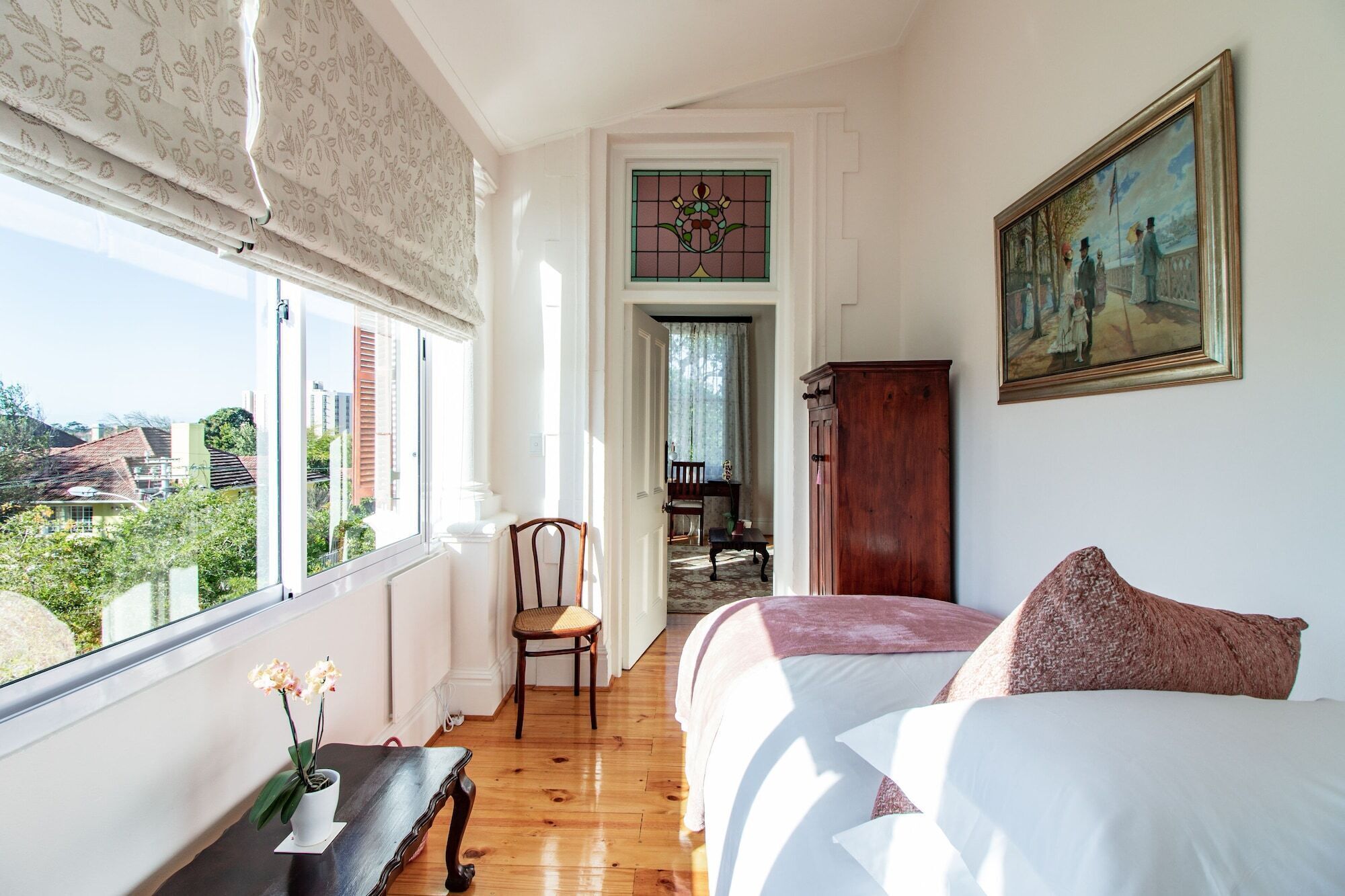 A bedroom with a large window, a bed, a chair, a wooden cabinet, and a painting on the wall.