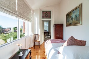 A bedroom with a large window, a bed, a chair, a wooden cabinet, and a painting on the wall. - Carmichael Guesthouse (Rosebank)