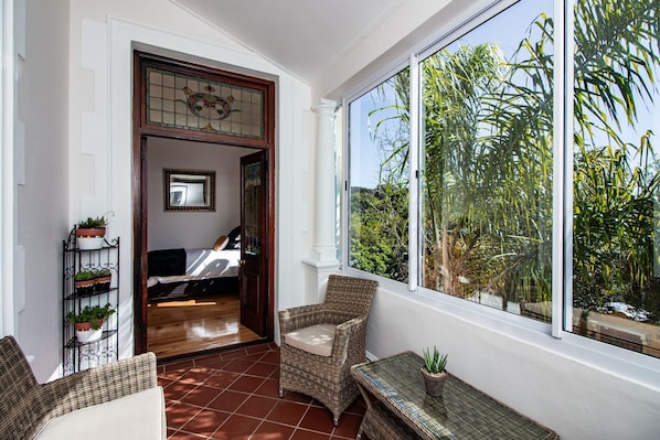 A sunlit room with wicker furniture, a large window with a view of greenery, and a door leading to another room. - Carmichael Guesthouse (Rosebank)