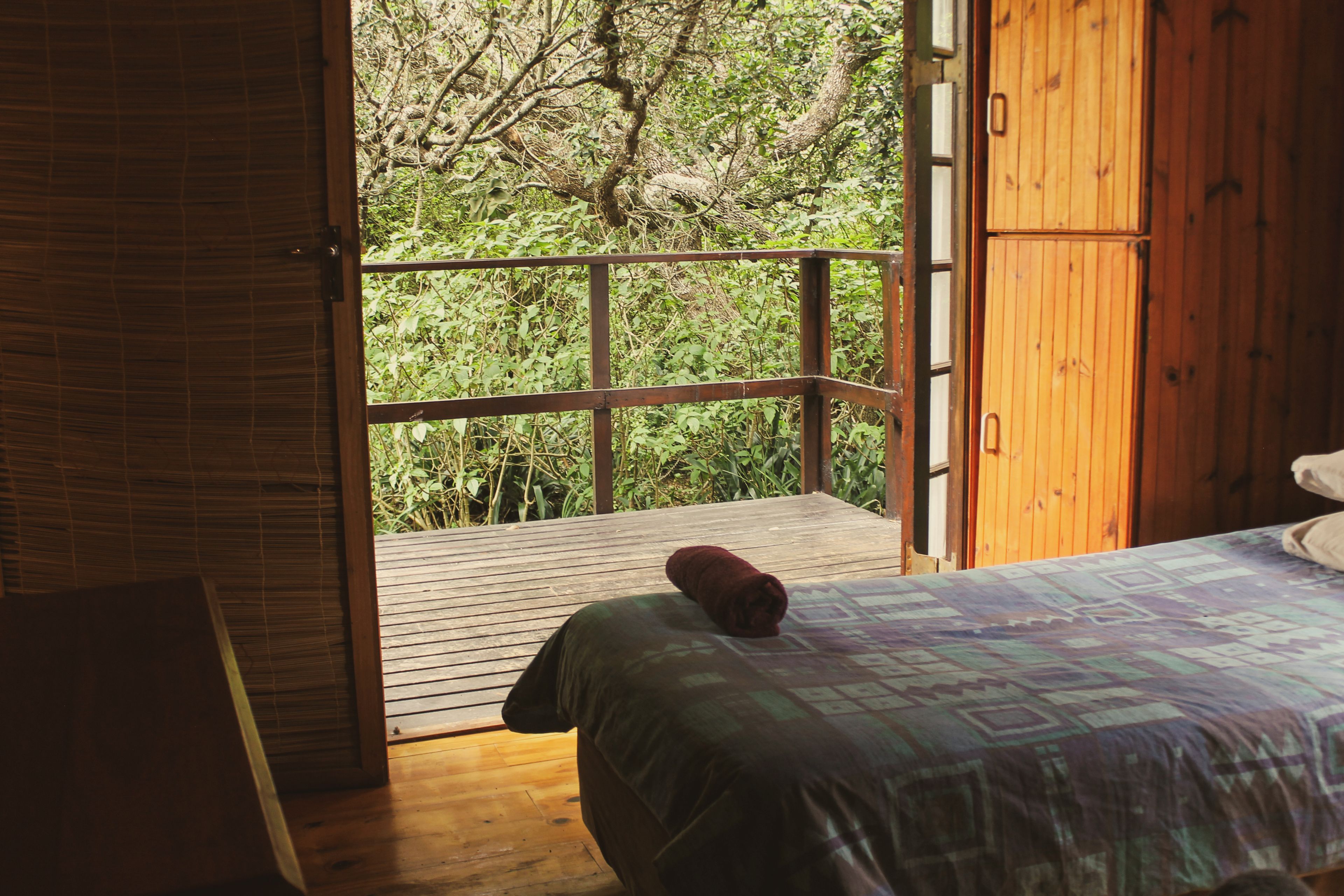 A room with a wooden floor, a bed with a patterned bedspread, and a view of greenery through an open door.