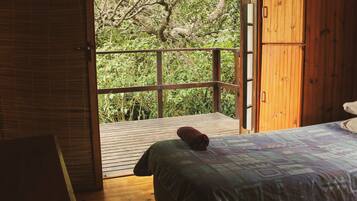 A room with a wooden floor, a bed with a patterned bedspread, and a view of greenery through an open door.
