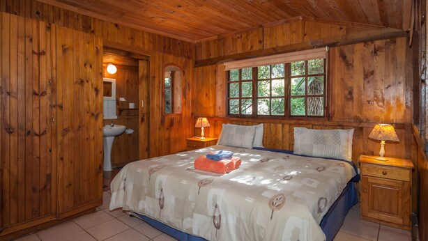 Un dormitorio de cabaña de madera con una cama grande, dos lámparas de noche, una ventana con vista a árboles y un armario de madera.