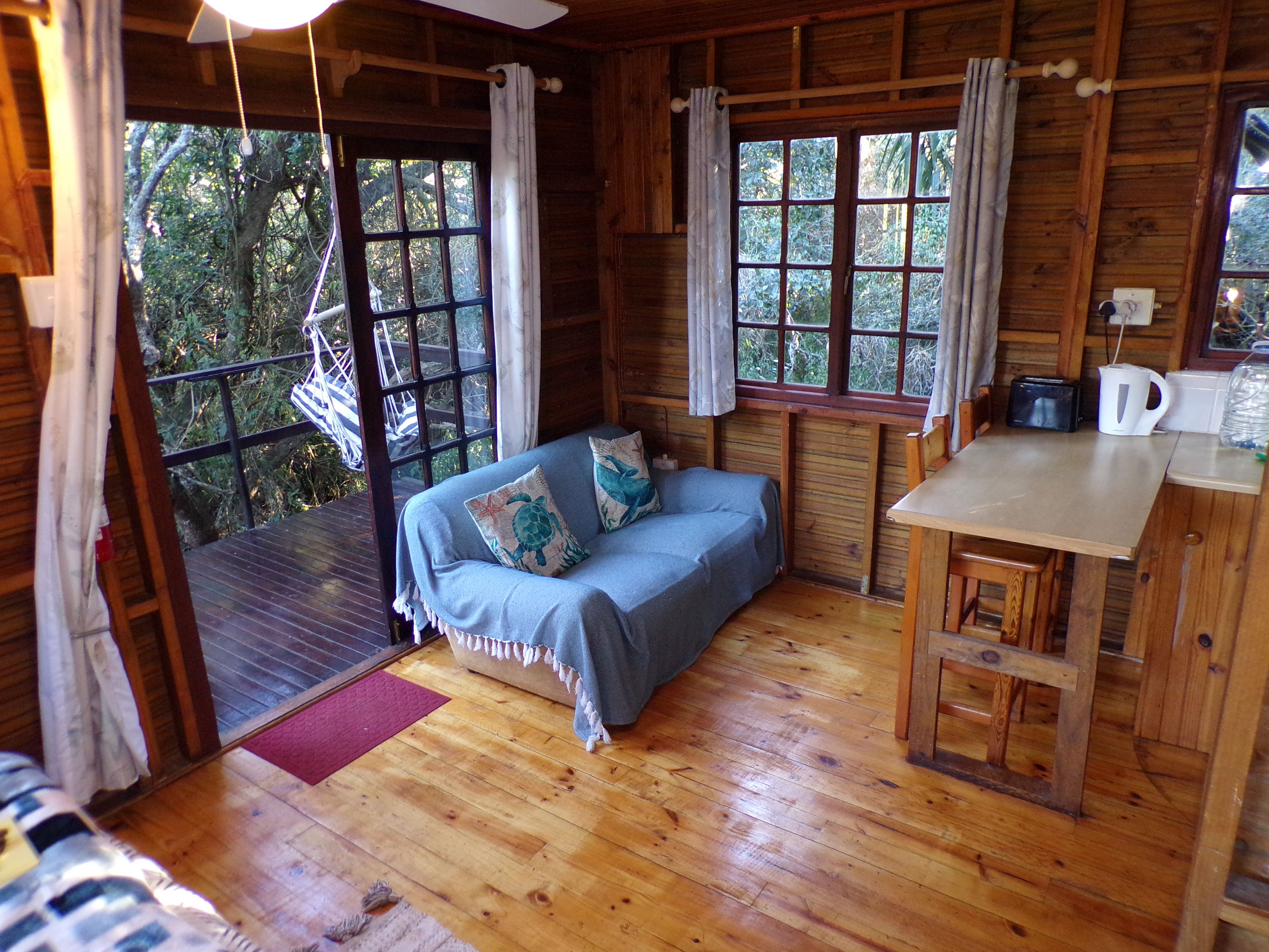 A cabin interior with a wooden floor, a sofa with pillows, a dining table, and a kitchen area.
