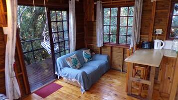 A cabin interior with a wooden floor, a sofa with pillows, a dining table, and a kitchen area.