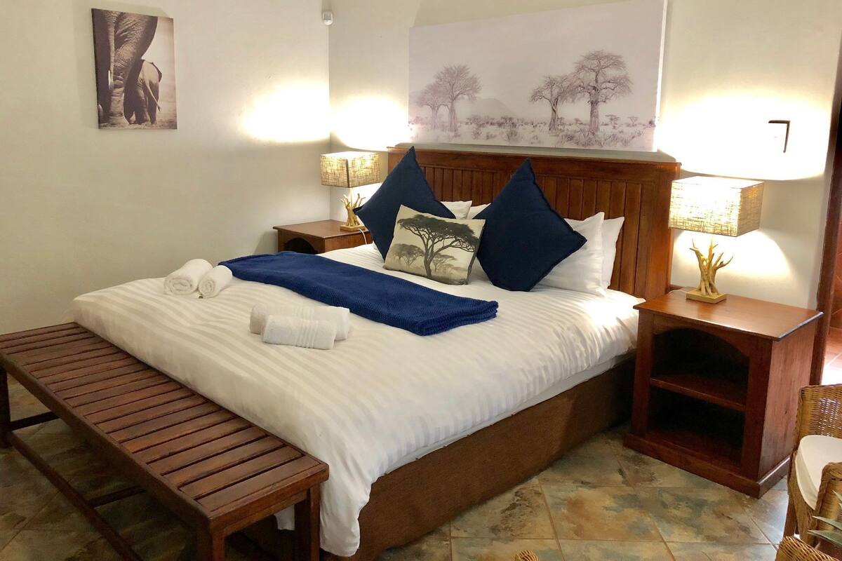 Una cama bien hecha con sábanas blancas, una manta azul y dos almohadas. Al pie de la cama, un banco de madera, una mesita de noche con una lámpara y un estante en la pared.