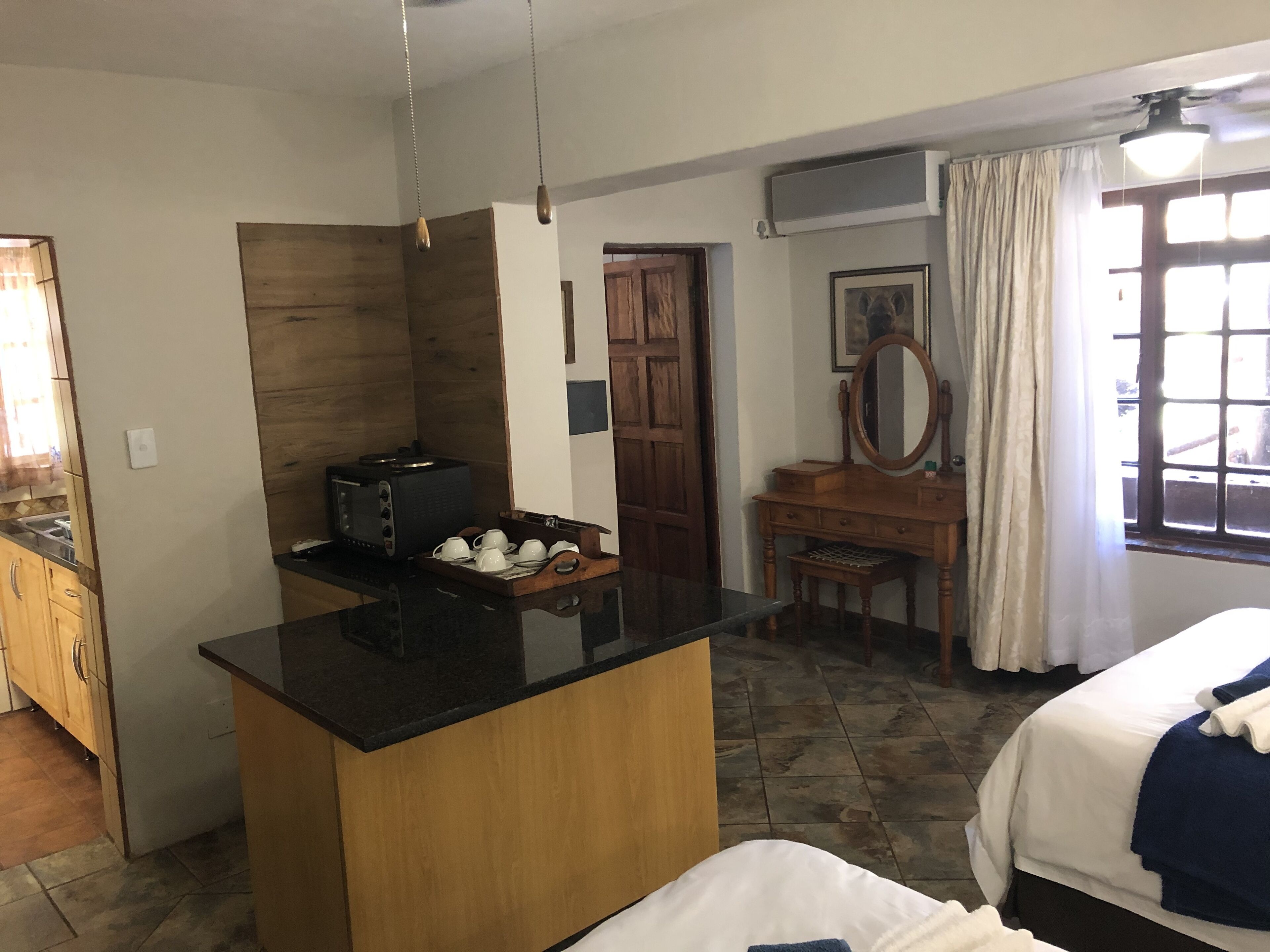 A hotel room with a kitchenette, a wooden dresser, a bed with a blue blanket, and a window with curtains.