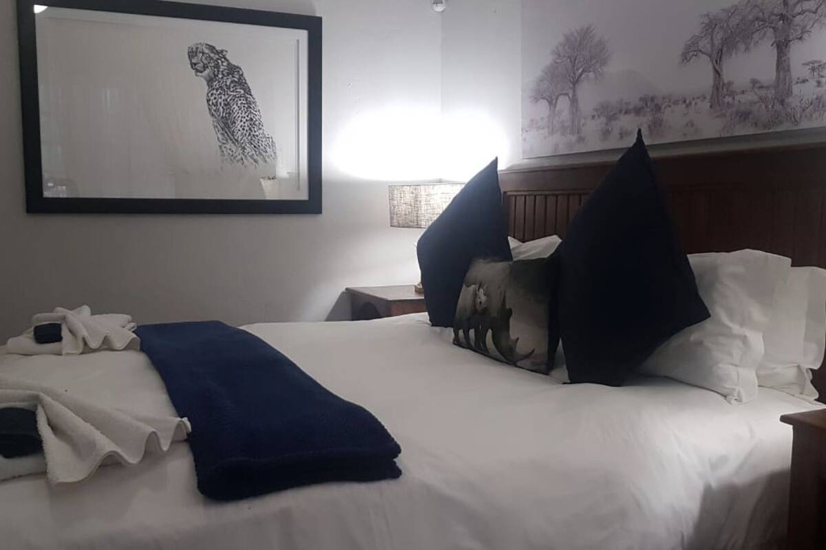 Una cama bien tendida con ropa de cama blanca y azul, un cuadro enmarcado de un guepardo en la pared, y una pintura de un paisaje sobre la cama.