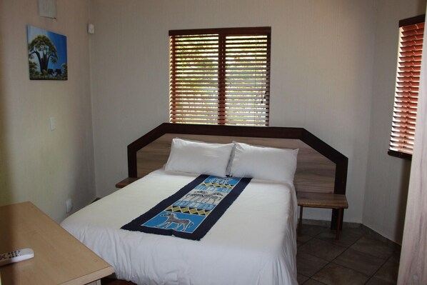 A bedroom with a bed, a wooden headboard, a window with blinds, a bedside table, and a wall-mounted painting.